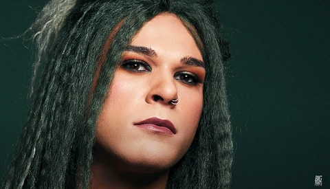 Portrait of a person with long dark hair against a teal background, with defined eye makeup and neutral expression.