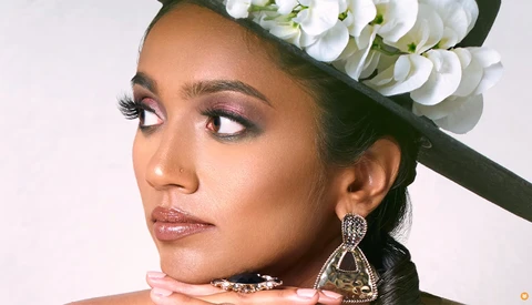 Portrait of a woman with elaborate eye makeup and white orchid headpiece, wearing statement earrings and looking to the side.