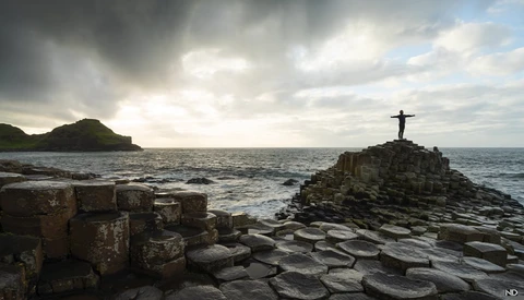 Figure standing on a rocky outcrop with a cross silhouetted against a dramatic cloudy seascape.