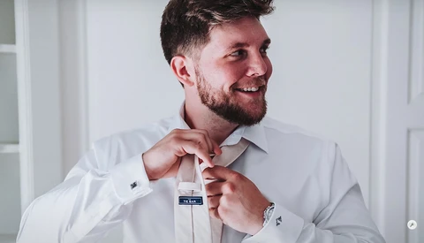 Man in white dress shirt adjusting tie with both hands while smiling.