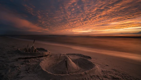 Sandcastle on a beach at sunset with dramatic orange and amber clouds overhead.