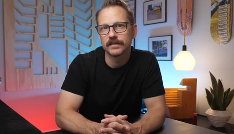 Man with glasses and mustache in black t-shirt seated at desk with studio lighting setup visible behind him.