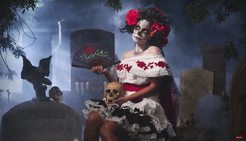 Woman in skull makeup and floral crown holding a hat in a cemetery at dusk.