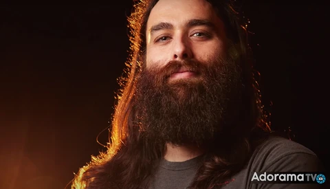 Man with long dark beard and hair illuminated by warm golden-orange backlighting against a dark background.