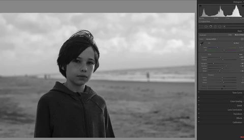 Young person in dark hoodie gazing toward horizon over calm water in black and white.