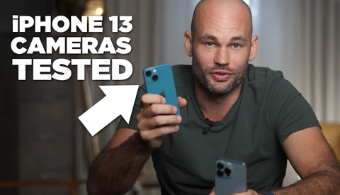 Man in dark t-shirt holding blue iPhone 13 toward camera with second device visible on table.