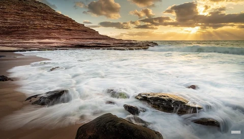 Waves roll onto a rocky beach at sunset, with layered cliff formations rising in the background.