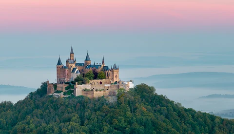 Medieval castle with multiple spires perched atop a forested hilltop at sunrise.
