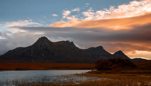 Mountain silhouetted against dramatic orange and blue sunset sky, reflected in still water below.