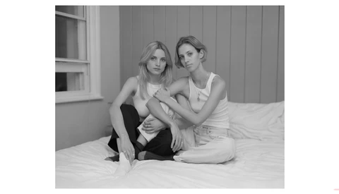 Two women sitting together on a bed, smiling at the camera in a black and white portrait.