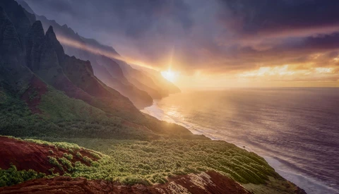Dramatic coastal landscape with steep green cliffs overlooking a beach and ocean at golden hour sunrise or sunset.
