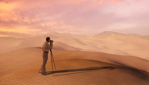 Photographer with tripod-mounted camera silhouetted against golden sand dunes at sunset.