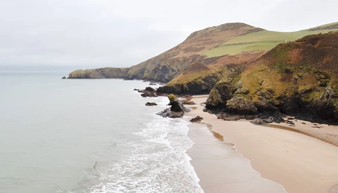 Coastal landscape with golden sand beach, rocky outcrops, and green hillsides meeting calm sea.