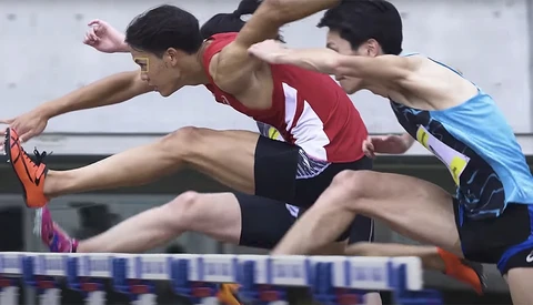 Three male athletes competing in a hurdles race, captured mid-stride as they approach a blue hurdle.