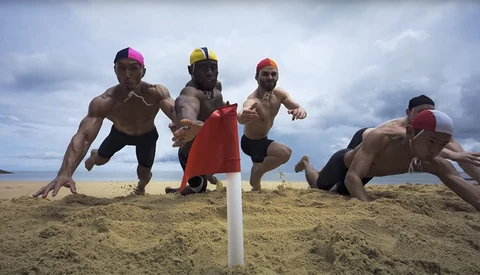 Five shirtless men in colored caps doing push-ups on sand with a white pole marker and cloudy sky behind them.