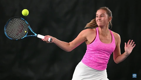 Female tennis player in mid-forehand stroke wearing pink tank top and white skirt against dark background.
