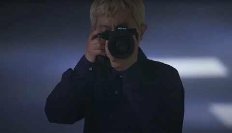Person holding a Nikon DSLR camera up to their face in a dark studio setting with blue-tinted lighting.