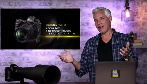 Man in plaid shirt gesturing while presenting camera equipment and specifications on screen.