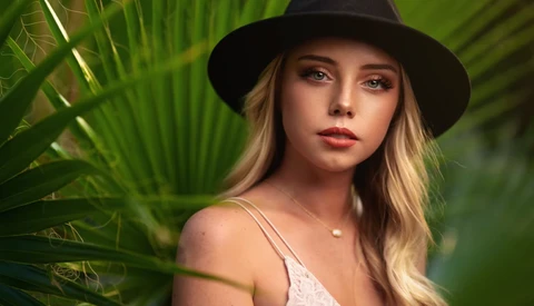 Woman wearing a black cowboy hat and white tank top, posed among green palm fronds.