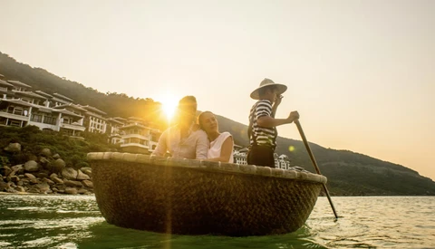 Family in a traditional woven boat on calm water during golden hour with hillside buildings in background.
