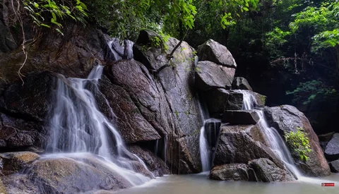 Multi-tiered waterfall cascading over large boulders surrounded by dense green forest.