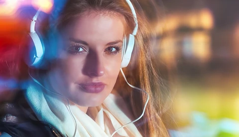 Woman wearing white headphones looking at camera with colorful bokeh lights in background.