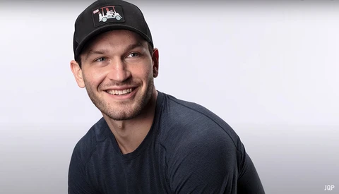 Smiling man wearing a black baseball cap and dark blue t-shirt against a neutral gray backdrop.
