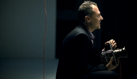 Man in dark clothing holding a film camera, photographed in profile against a minimalist dark background.