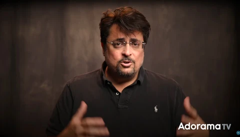 Man in black polo shirt gesturing while speaking against neutral backdrop.