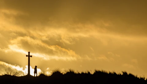 Silhouetted figure standing beside a cross on a hilltop against a golden sunset sky.