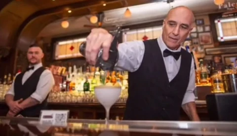 Bartender in formal attire pouring an ingredient into a cocktail glass behind an upscale bar counter.