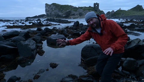 Photographer in red jacket reaching toward tide pool rocks during golden hour coastal shoot.