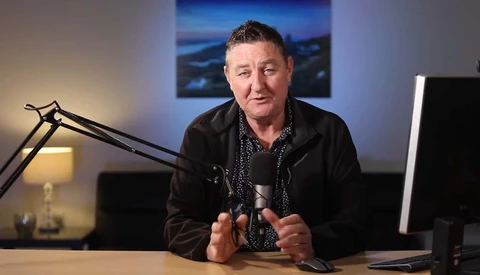 Man speaking into microphone at desk in professional studio setting with monitor and landscape photograph visible in background.