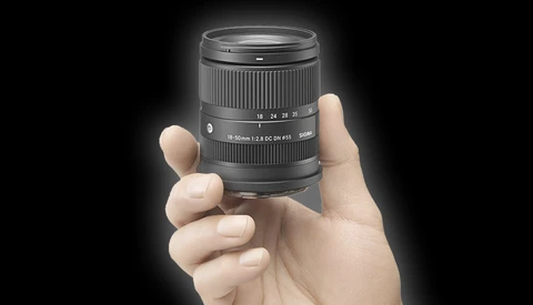 Hand holding a compact telephoto zoom lens with visible focal length markings and aperture information.