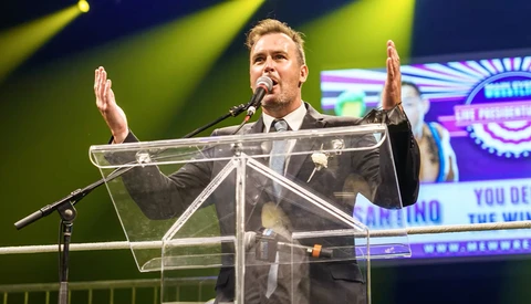 Man in business suit speaking at transparent podium with microphone, hands raised expressively.