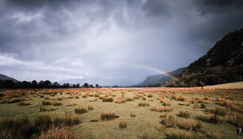 Expansive grassland valley beneath dramatic storm clouds, with forested mountains flanking both sides.