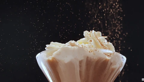 Creamy beverage in glass cup with splashing liquid and particles suspended in air against dark background.
