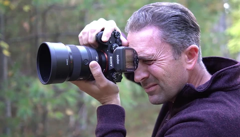 Male photographer holding a mirrorless camera with telephoto lens to his eye, focusing intently on a subject outdoors.