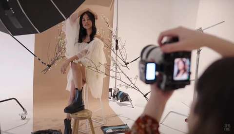 Behind-the-scenes view of a studio portrait shoot with model on stool and photographer operating camera in foreground.