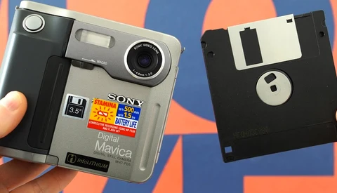 Sony Cyber-shot digital camera next to a floppy disk against a colorful striped background.