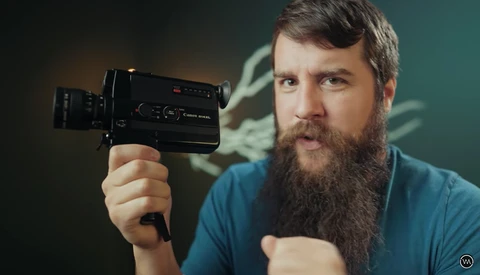 Bearded man in blue shirt holding a vintage film camera against a teal background.