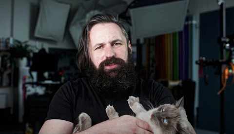 Bearded man holding a small tabby kitten in a studio setting with professional lighting equipment visible in background.