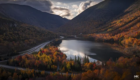 Mountain lake surrounded by autumn forest with road winding through golden and orange trees under dramatic sky.