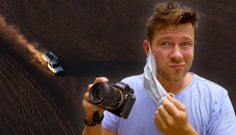 Photographer holding a professional camera with a towel around his neck, aerial view of a car on a desert road in background.
