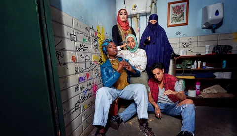 Six people of varying ages posing together in a modest room with blue walls and graffiti-covered surfaces.