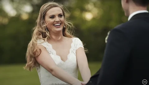 Bride in white lace wedding dress smiling at groom during outdoor ceremony.