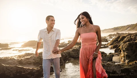 Couple holding hands walking along rocky coastline at golden hour sunset.