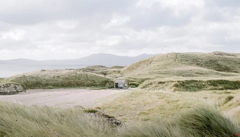 Coastal dune landscape with sparse vegetation, a small structure, and mountains across the water under overcast sky.