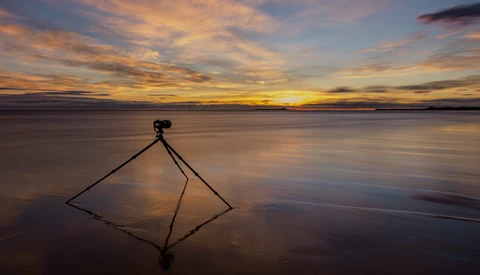 Camera on tripod positioned in shallow water during golden hour sunset with mirror reflection.