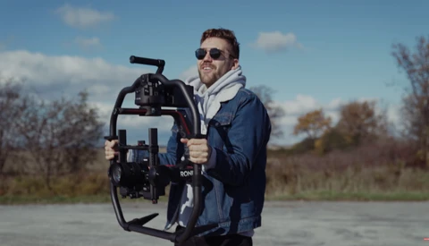 Videographer holding a cinema camera rig outdoors on a paved surface with bare trees in background.